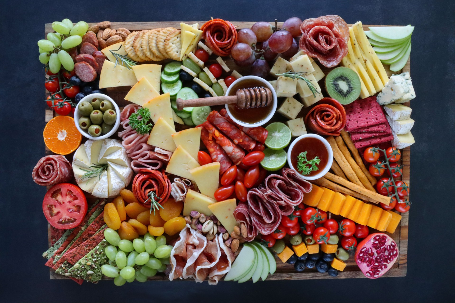 Close-up image charcuterie board ingredients, crackers, ham, prosciutto and salami roses, cucumber, lime half, cherry tomatoes, Brie, kiwi, Cheddar, red grapes, ramekin of honey and honey dipper, black background, elevated view